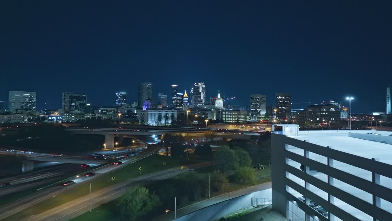 Car park with busy highway at night in Atlanta City, Georgia. Aerial forward wide shot. Skyline and skyscrapers in distance. Lighting buildings of downtown.