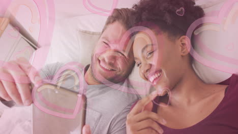 animation of hearts over diverse couple using tablet on bed