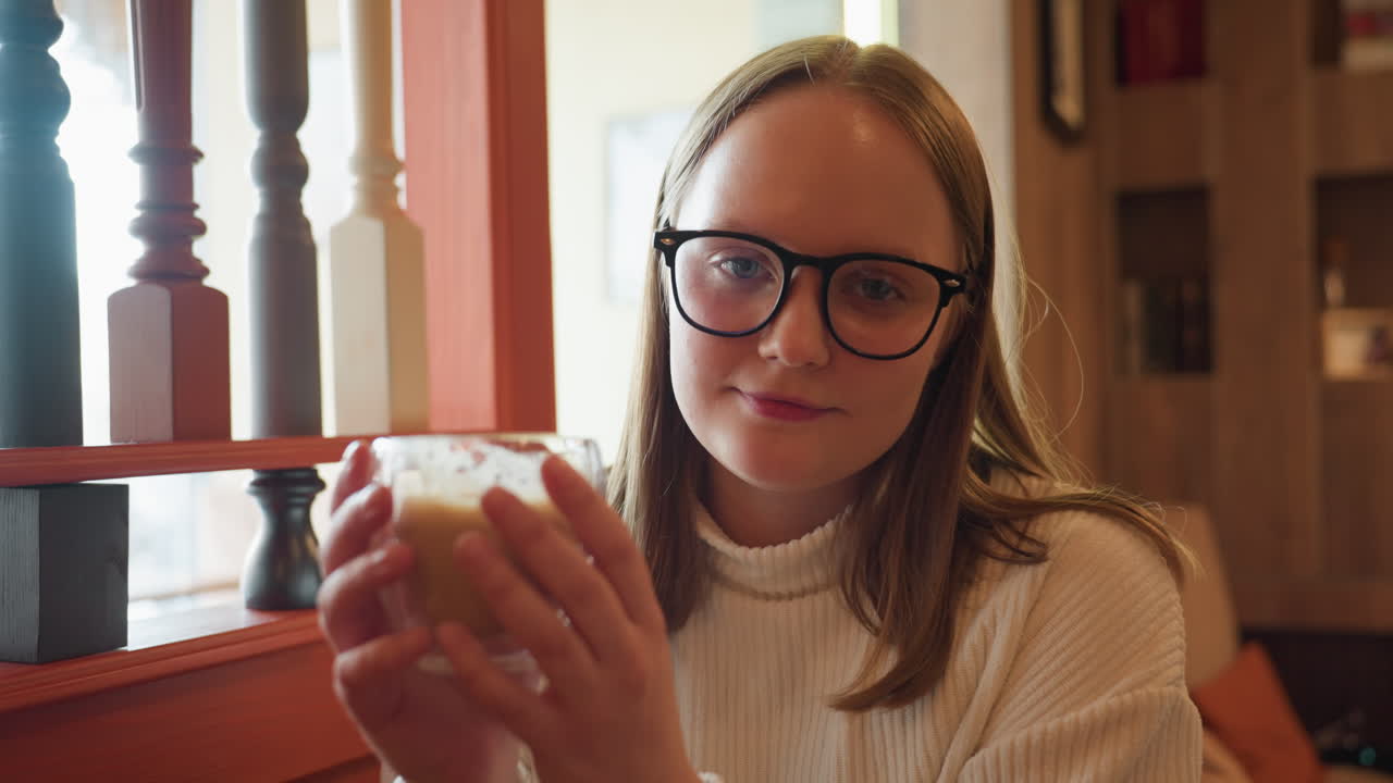 young woman in white sweater with glasses holds coffee cup while gazing calmly at camera, soft lighting highlights her serene expression, blurred warm background with wooden cabinet