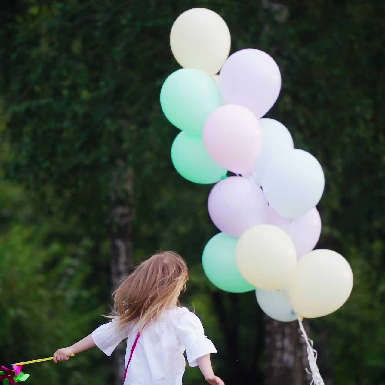 Back view of a little girl in the park. Small girl in blue jeans runs to the lots of air balloons on the natural green background in summer.