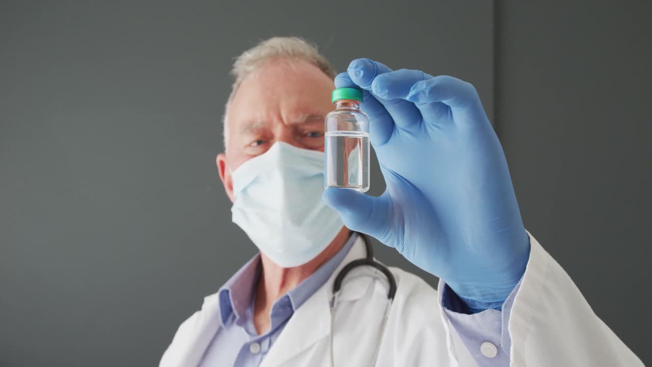 Ritratto di medico anziano caucasico con mascherina che tiene in mano il vaccino COVID-19.