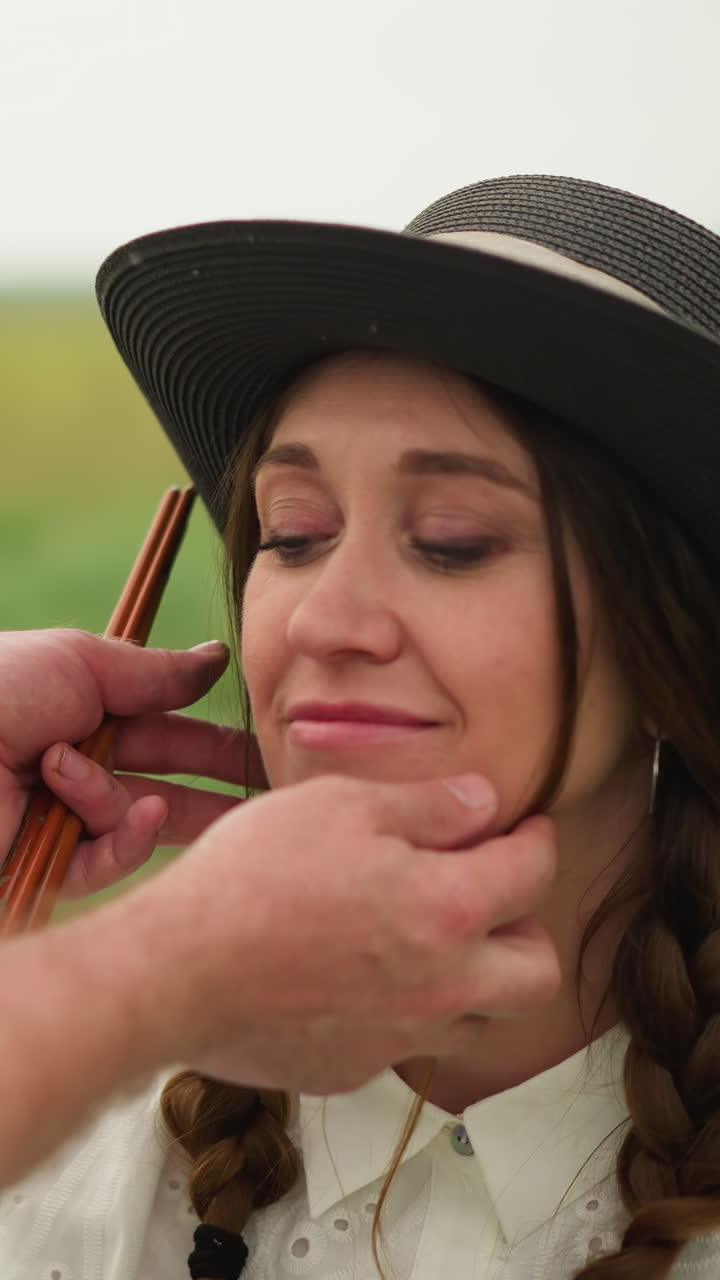 un artesano con una camisa a cuadros ajusta suavemente el sombrero de una mujer con un vestido blanco, su cabello trenzado y su sonrisa iluminan el sereno campo verde