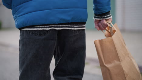 back view of individual's arm and side in bright blue puffer jacket holding a shopping bag, set against a blurred urban background, highlighting everyday casual life