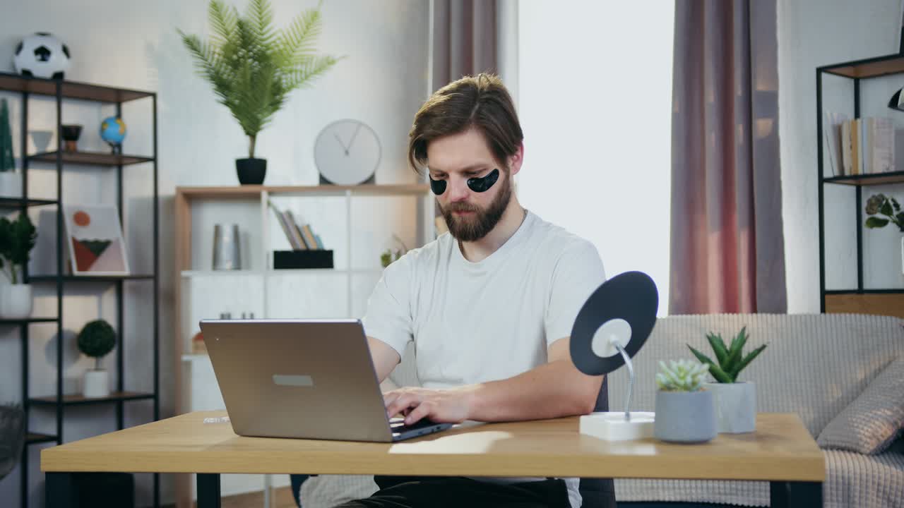 Good-looking smiling modern 30-aged bearder with anti-aging hydrogel eye-patches sitting at the table and working on computer,close up