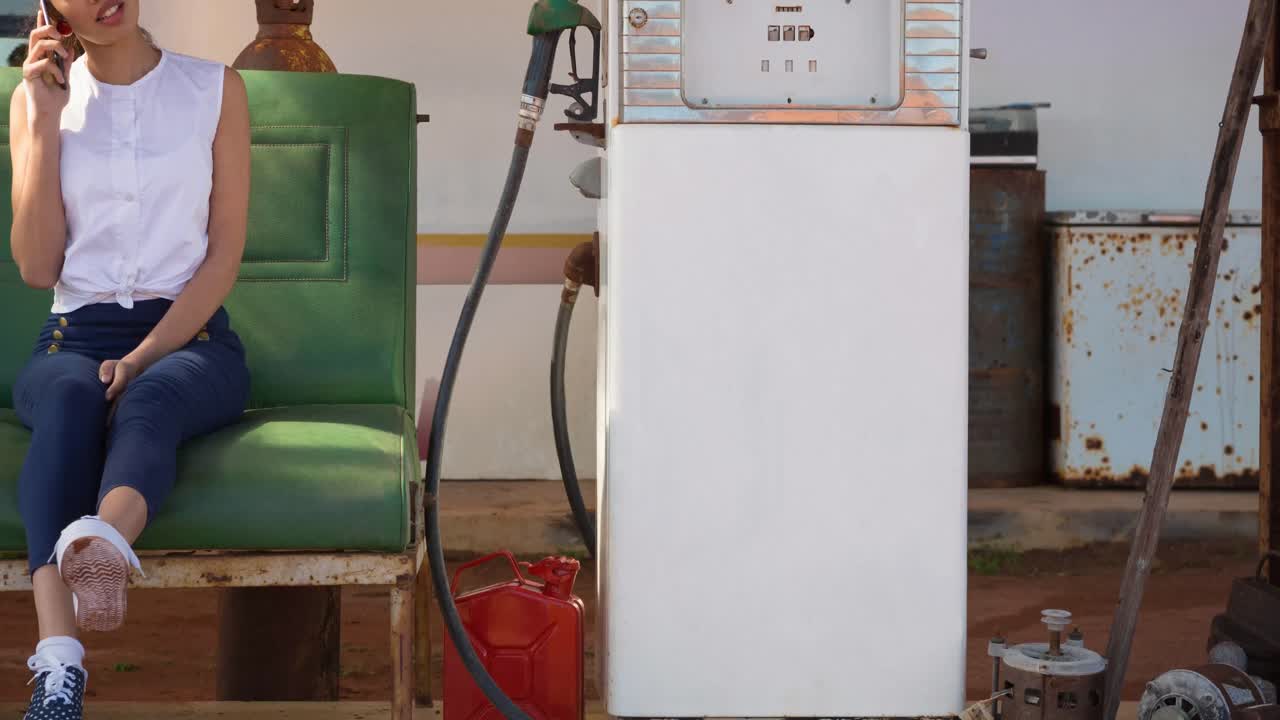 mujer biracial hablando en un teléfono inteligente sentada junto a una bomba de combustible en una gasolinera