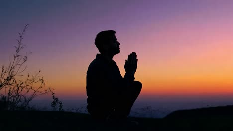 silhouette of a man squatting and praying at sunset, surrounded by a vibrant sky filled with orange and purple hues