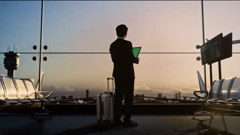 full body back view of asian businessman with rolling suitcase in boarding lounge at the airport, using green screen tablet while waiting for flight, airplane takes off outside the window