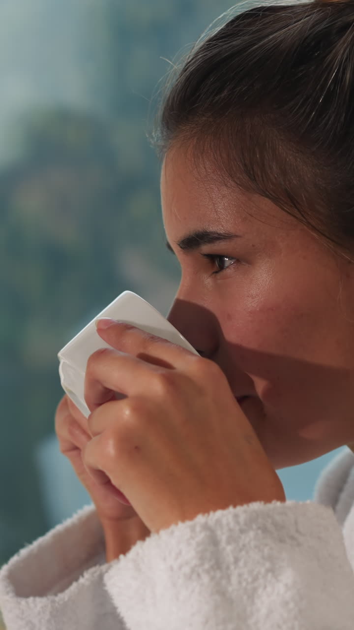 una mujer bebe café cerca de la ventana. una mujer muy positiva con túnica de terry toma un sorbo de té natural recién preparado en la cocina de casa. estilo de vida saludable