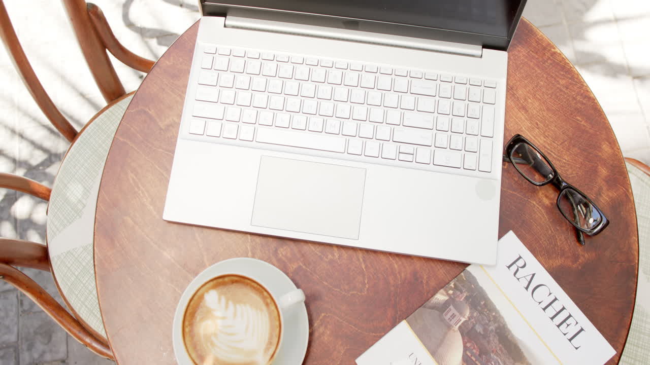 A laptop is open on a round table next to a cup of coffee and eyeglasses in a cafe