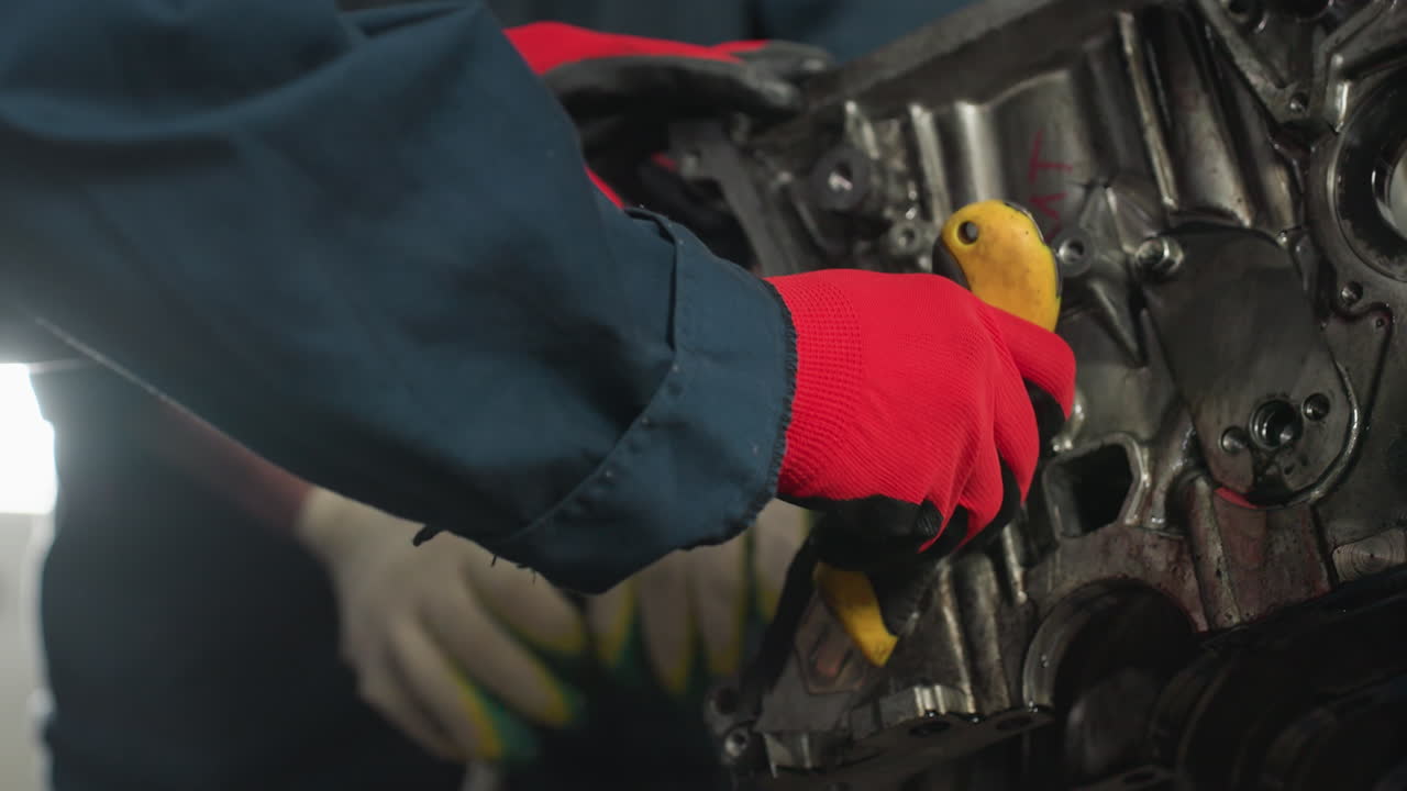 primer plano de un mecánico en uniforme azul usando un raspador amarillo en un motor de coche, otro mecánico con guantes rojos ayuda, en un taller automotriz ocupado