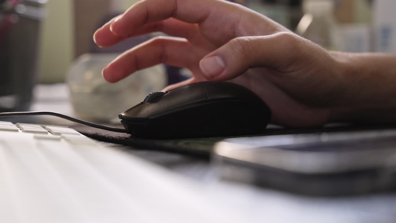 a hand using a wired computer mouse on a desk in a bright office. Features a white keyboard, shallow depth of field, and a professional workspace setting for technology themes