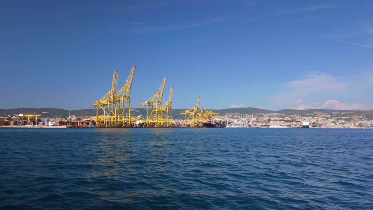 Gru da carico gialle si ergono lungo una banchina portuale trafficata a Trieste, Italia, viste attraverso l'acqua calma del mare blu sotto un cielo sereno e soleggiato