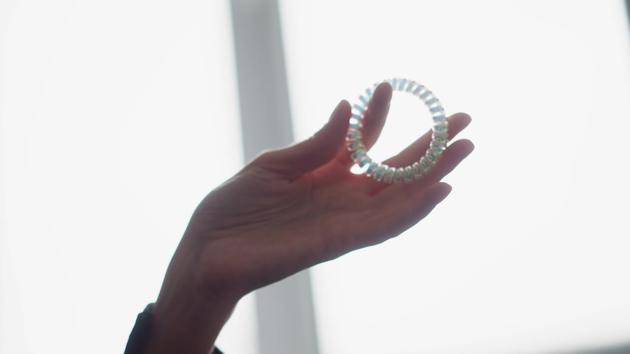 Primo piano di una mano che tiene e ruota delicatamente un elastico per capelli iridescente contro la luce brillante di una finestra. L'attenzione rimane sui dettagli della texture, sugli effetti di luce soffusa e sul sottile movimento delle dita.