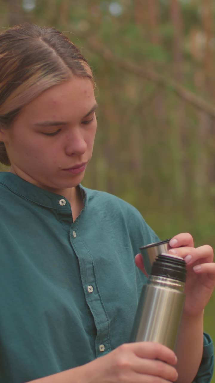 vista lateral de las hermanas pausa en el bosque tranquilo, una abre el termo presionando la cubierta interior, mientras que la otra cepilla suavemente su cabello, las mochilas descansan en el árbol caído mientras disfrutan de un momento sereno juntos
