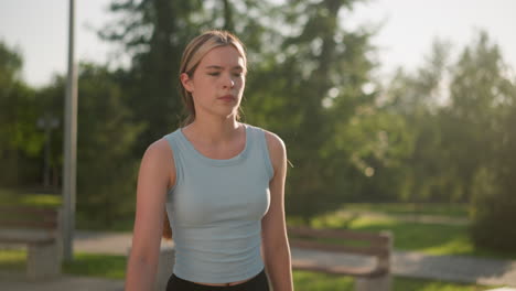 lady wearing cyan top looking thoughtful while moving as if skating, sunlight reflecting off her, creating soft backlight effect, blurred background features greenery, benches and lamp posts