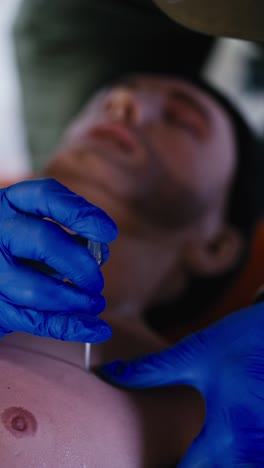 video vertical de primer plano de un médico masculino con guantes elásticos azules en una muñeca masculina practicando la inserción de una aguja médica en el pecho de un hombre durante un curso militar médico