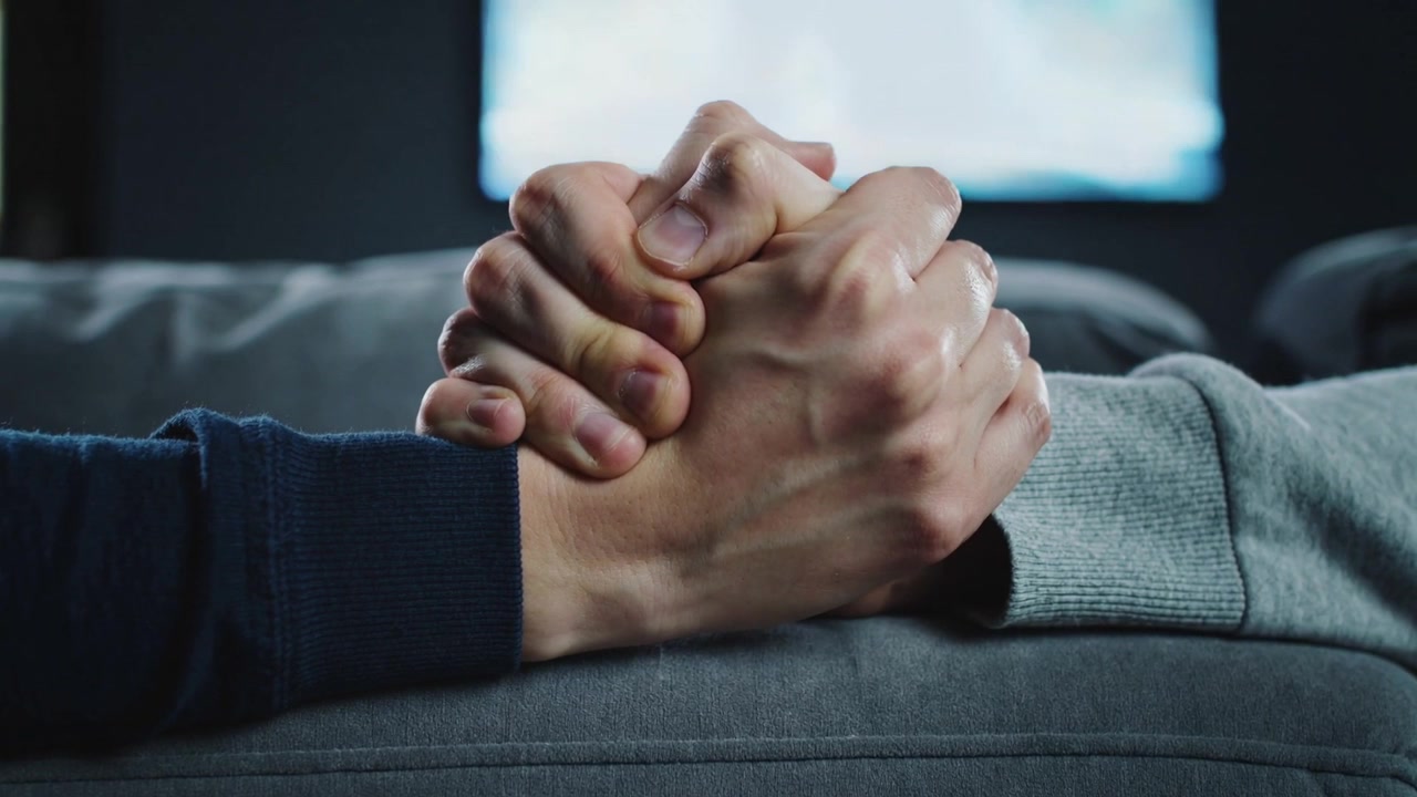 Holding two adult hands fingers weaving on couch armrest after touch, navy grey sleeves, blurred TV