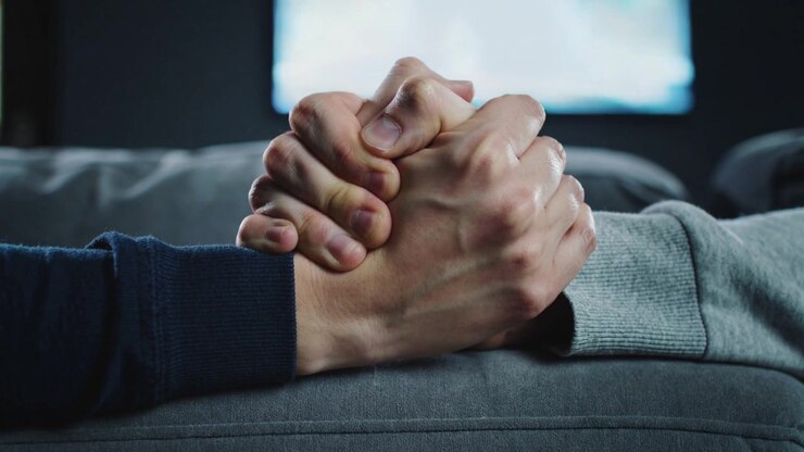 Duas mãos adultas com dedos entrelaçados no braço do sofá após um toque, mangas azul-marinho e cinza, e TV desfocada.