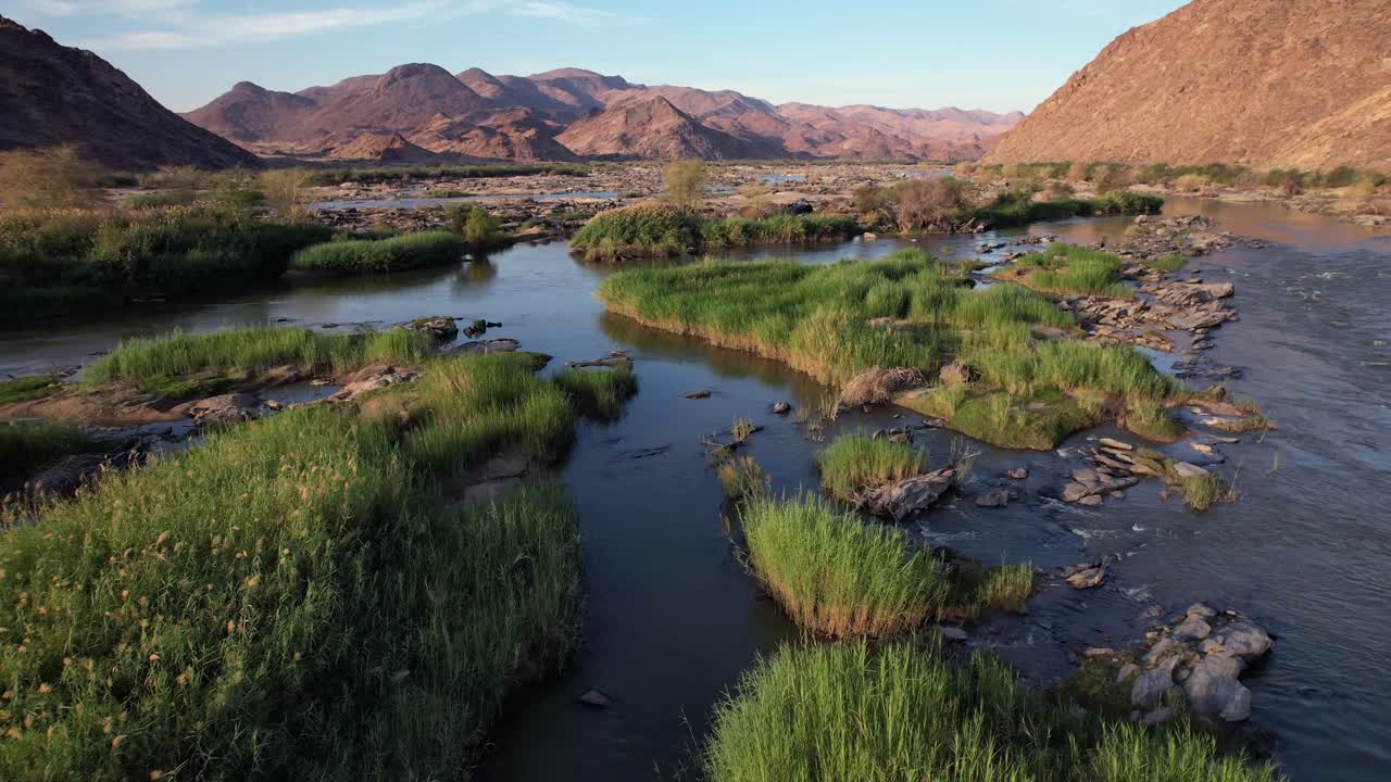 Drone flies backward and ascends over a river dotted with green grassy islands, revealing mountains bathed in warm golden hour light. Scenic natural landscape in motion