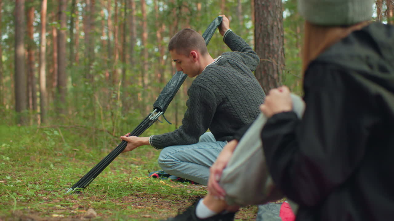 Close up rear view of lady seated watching boyfriend squat in forest while he carefully brings out and holds tent pole during calm camping preparation among trees and greenery