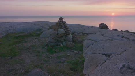 Una-Escultura-De-Equilibrio-De-Rocas-Construida-En-La-Cima-De-Una-Montaña,-Con-Vista-Al-Océano-En-Calma-Con-Una-Hermosa-Puesta-De-Sol-Rosa-Y-Un-Cielo-Despejado