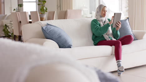 Biracial-woman-in-hijab-using-tablet-on-sofa-at-home-with-copy-space,-slow-motion