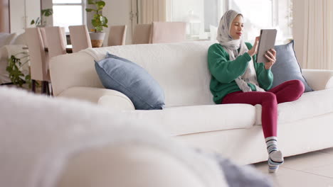 mujer biracial en hijab usando tableta en el sofá en casa con espacio de copia, cámara lenta