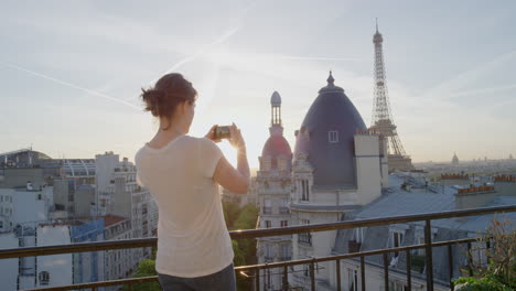 Mujer-Joven-Usando-Un-Teléfono-Inteligente-Tomando-Fotos-Disfrutando-Compartiendo-Experiencias-De-Vacaciones-De-Verano-En-París-Fotografiando-La-Hermosa-Vista-Del-Atardecer-De-La-Torre-Eiffel-En-El-Balcón