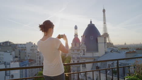 young woman using smartphone taking photo enjoying sharing summer vacation experience in paris photographing beautiful sunset view of eiffel tower on balcony