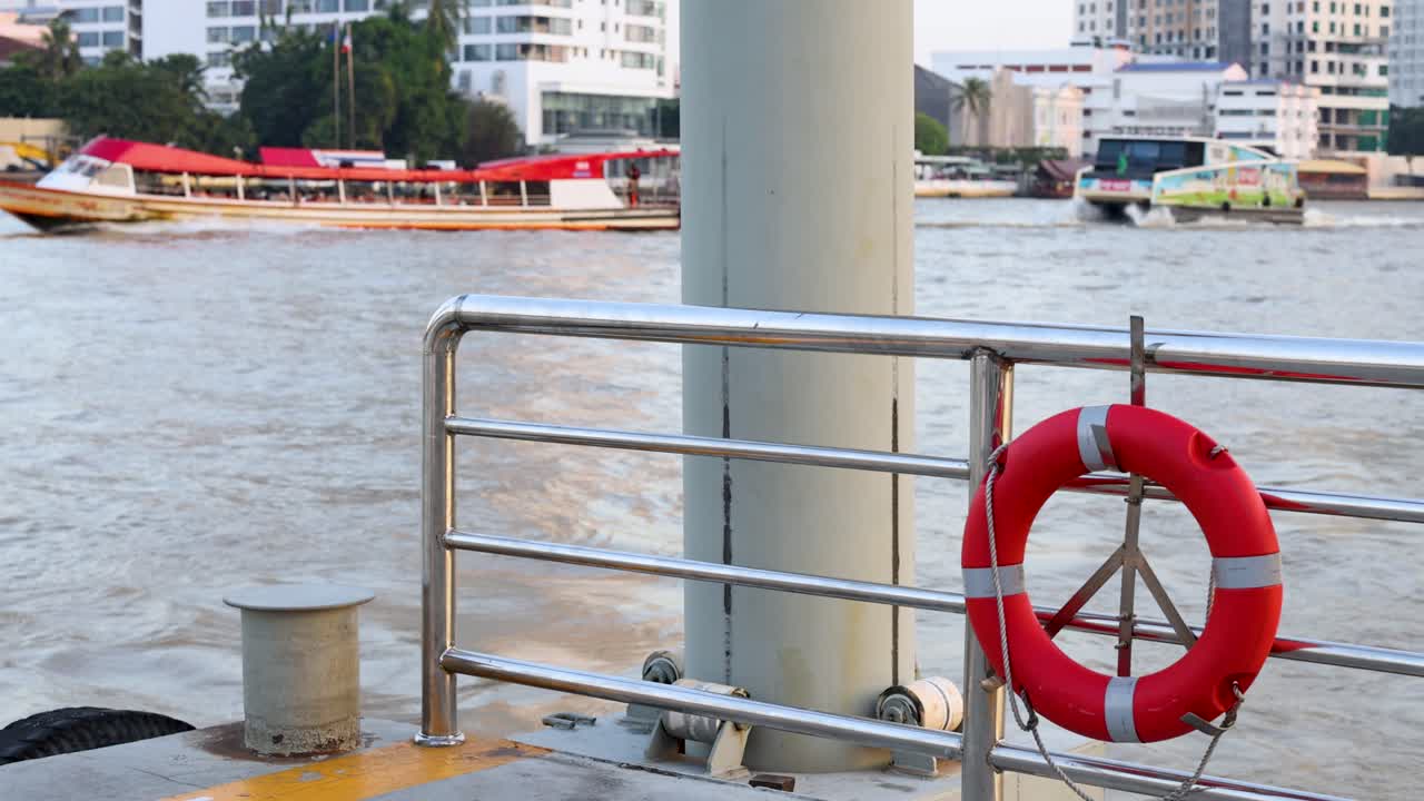 Static pier view with red lifebuoy as passenger boats cruise along urban Bangkok riverfront