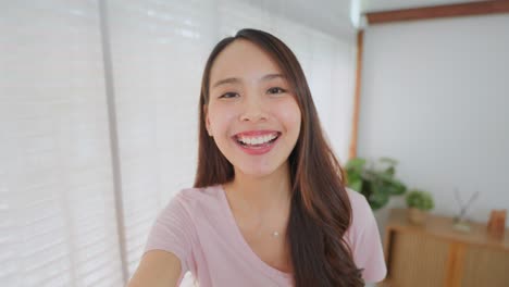 Point-of-view-vlog-or-selfie-camera-of-Asian-beautiful-happy-woman-blogger-holding-phone-looking-at-camera,-greeting-and-talking-to-friends-or-family-on-video-call-online.-Technology-lifestyle-concept