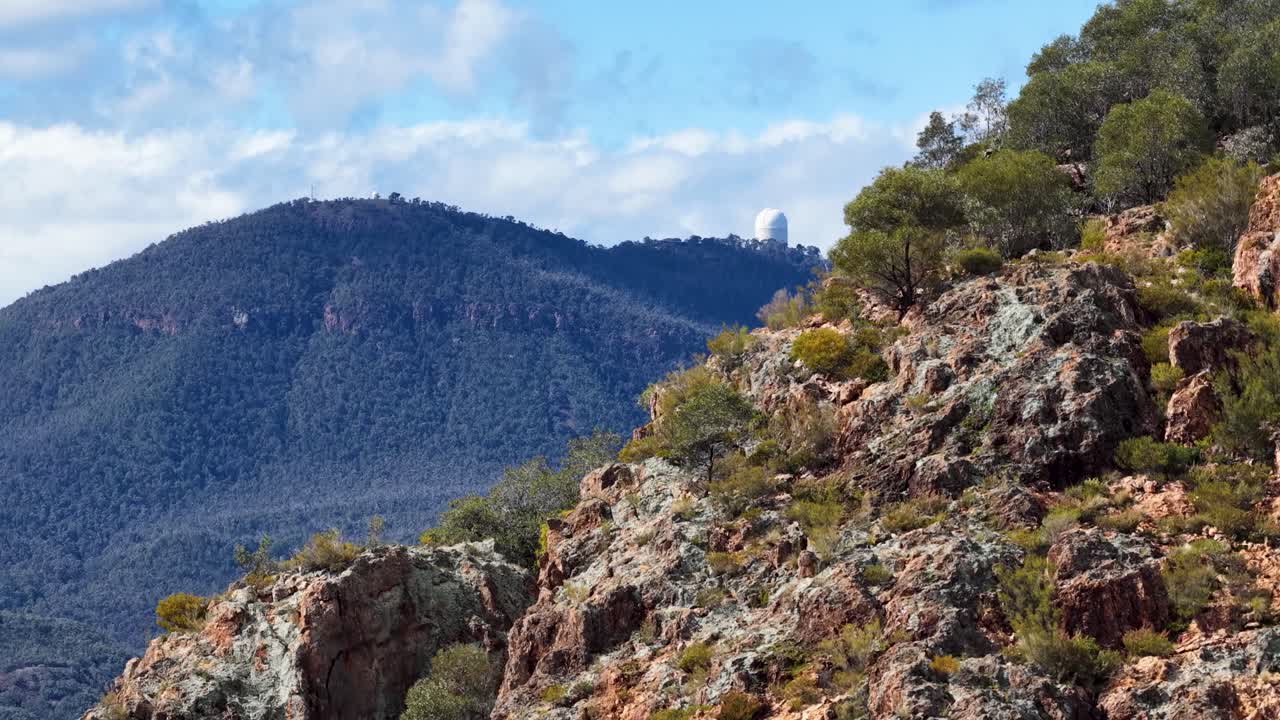 Camera slowly pans right across rugged, sunlit hills with sparse trees, revealing a distant observatory dome atop a forested mountain under blue sky