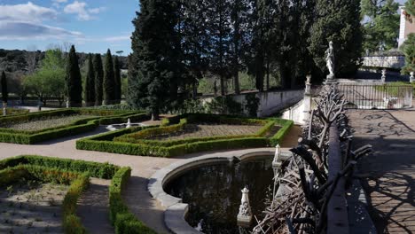 panning to the left general shot of a baroque garden, with fountains, and a fence with dead branches in the right at the beginning