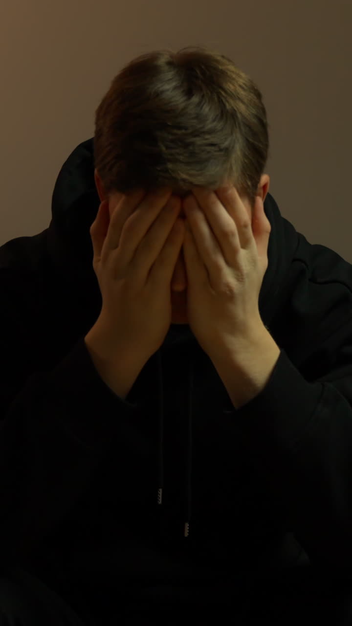 Young man in a black hoodie covers his mouth with both hands, staring in shock and worry under warm indoor lighting—concept of stress, panic, and personal crisis