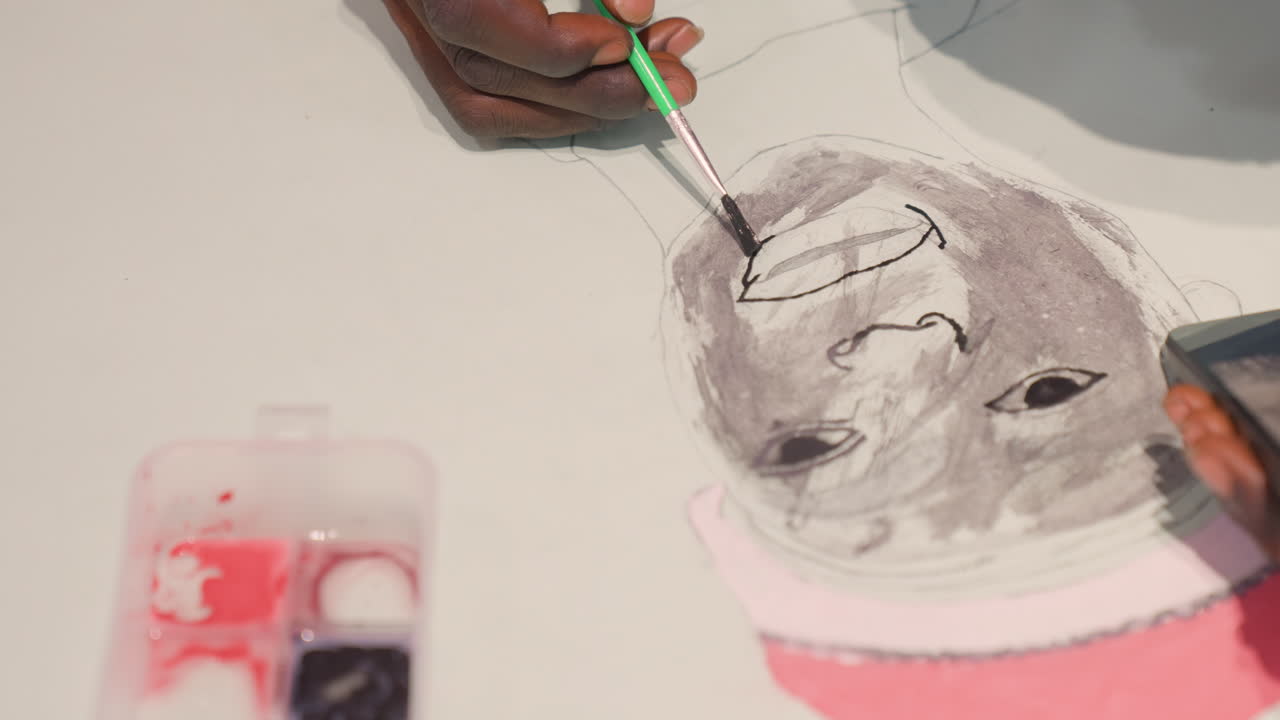 Closeup black artist adding finishing strokes to lips, green brush shaping small mouth detail on monochrome portrait, pink hat accent and paint case nearby, warm studio mood with careful playful touch