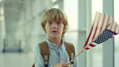 Close-Up-Of-The-Cute-Small-Boy-Standing-In-The-Airport-Passage,-Looking-To-The-Camera-And-Waving-With-American-Flag