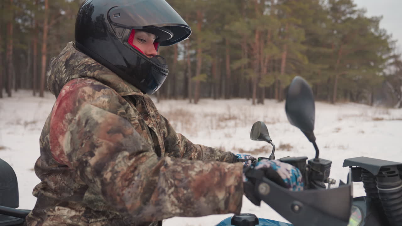 Un pilota mimetico su un quad sulla neve si prepara su un sentiero nella pineta, regolando lo specchietto del casco mentre stringe l'acceleratore. Primo piano sulla mano guantata sul manubrio e sullo specchietto. Luce fredda e nuvolosa, palette di colori tenui.