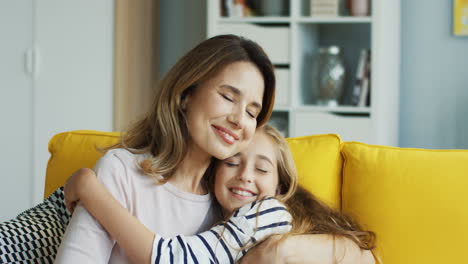 Madre-Feliz-Sentada-En-El-Sofá-Amarillo-Y-Sonriendo,-Luego-Su-Hija-Adolescente-Se-Acerca-Y-La-Abraza