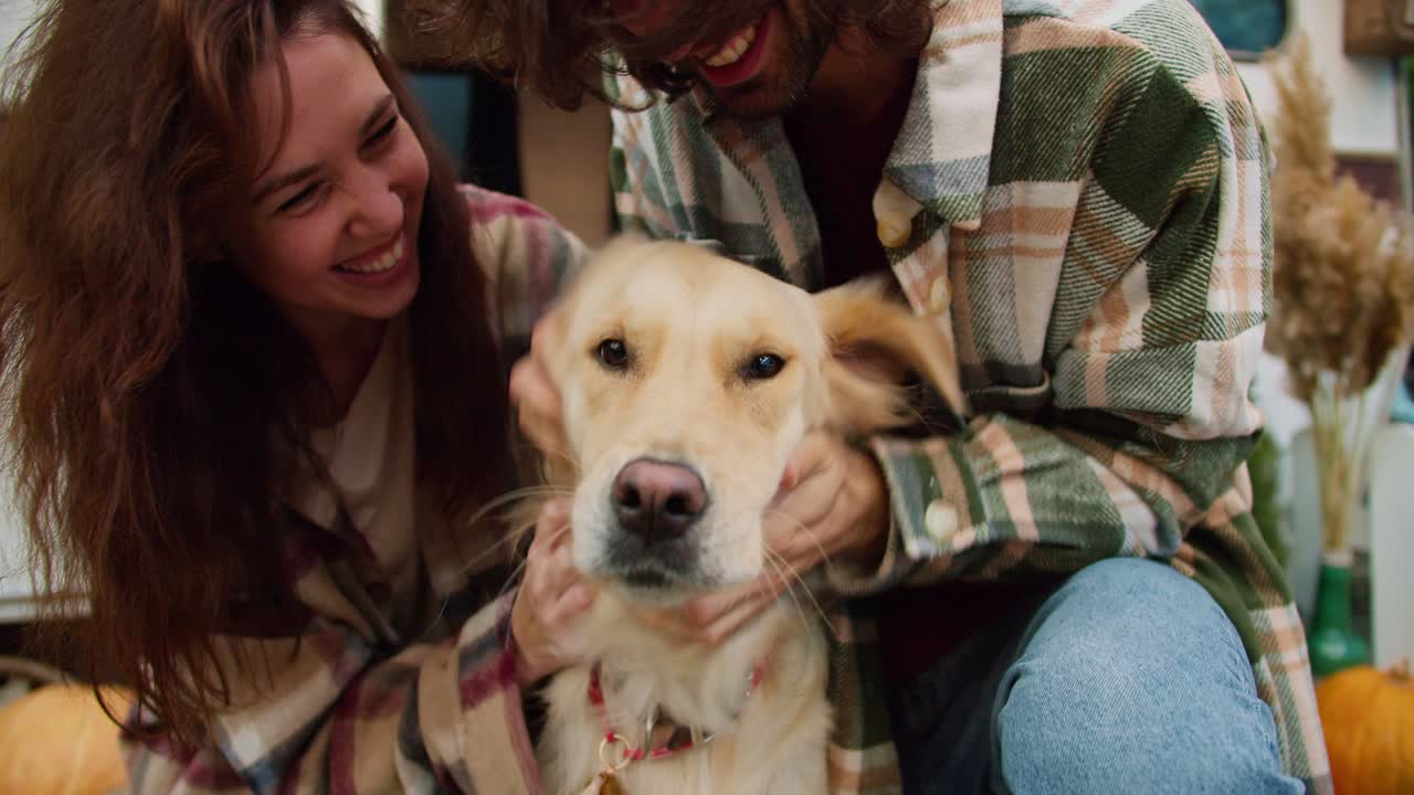 una foto en primer plano de una feliz chica morena con una camisa a cuadros junto con su novio con una camiseta a cuadros verde acariciando a su gran perro de color crema cerca de un remolque en un campamento durante un picnic fuera de la ciudad en verano