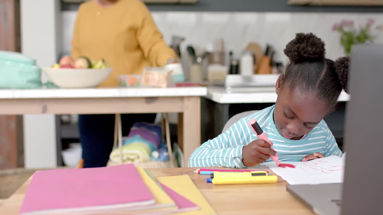 African american girl learning online at table using laptop with mother in kitchen, slow motion