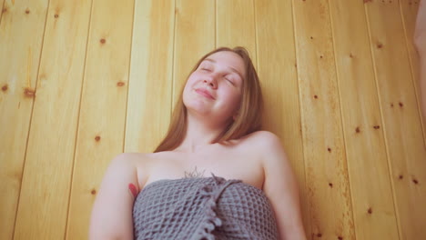 young housewife leans back peacefully against wooden wall with eyes closed, wrapped in textured towel, enjoying quiet restful moment, face lit by soft natural light, expressing calm relaxation indoors