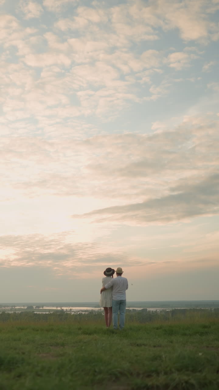 해가 지는 동안 호수 의 잔디 언덕에 서 있는 한 부부의 뒷면. 색 셔츠, 모자, 청바지를 입은 남자가 여자를 허리에 붙잡고 있다. 그녀는 검은 모자와 색 드레스를 입고 있다.