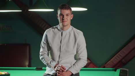 young man in white shirt and grey trousers sits on green pool table, holding cue stick and smiling. pool balls and billiards setup are visible in the background under warm lighting