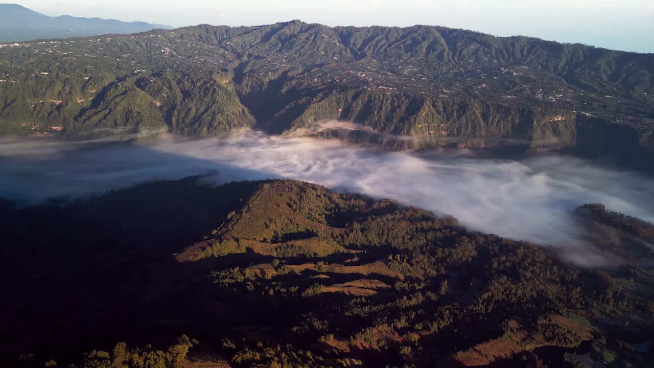 Paesaggio montano a Bali con colline boscose che si ergono sopra un mare di nuvole in una calda luce dorata, catturato da una prospettiva aerea con drone, creando una vista panoramica e suggestiva.