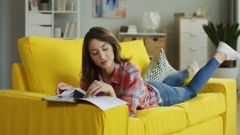 Mujer-Joven-Y-Guapa-Tendida-En-El-Sofá-Amarillo,-Pasando-Páginas-Y-Leyendo-Revistas-De-Moda-En-Casa