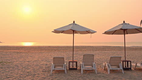 a couple of plastic seats and umbrellas on an empty white sand beach with golden sun over the ocean during sunset