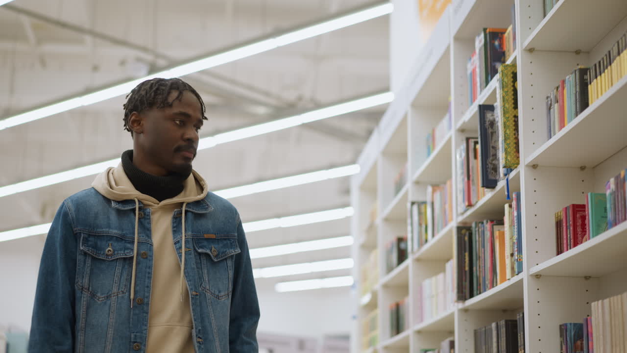 Young guy standing in bookstore, scanning colorful books on shelves. Bright indoor lighting highlights neat arrangement of books. Focus on casual exploration in modern bookstore environment