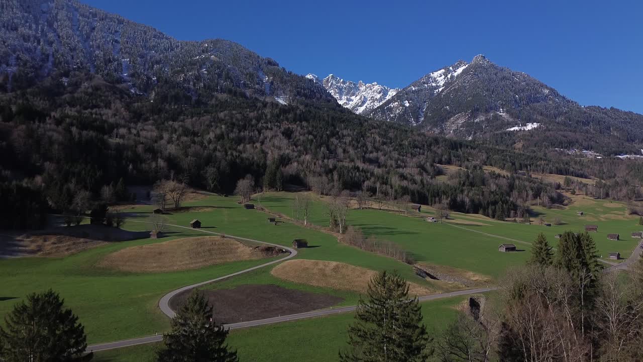 vista aérea de carreteras y pequeñas chozas con montañas cubiertas de nieve en el fondo en un hermoso día soleado con un cielo azul claro en austria