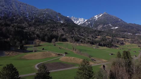 Aerial-view-of-road-and-small-huts-with-snow-capped-mountains-in-background-on-a-beautiful-sunny-day-with-clear-blue-sky-in-Austria