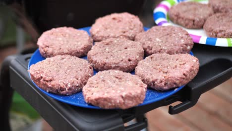 4K-Clip-In-Zeitlupe-Von-Ungekochten-Burgern-Mit-Alternativem-Fleisch-Auf-Bunten-Tellern-Neben-Dem-Grill,-Bereit-Zum-Kochen